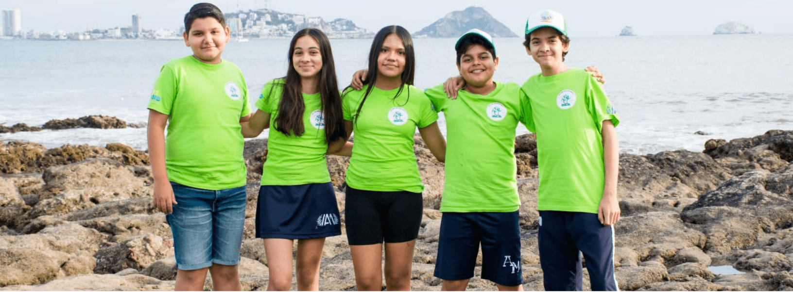 Alumnos del Instituto Anglo Moderno con playeras verdes frente al mar en Mazatlán